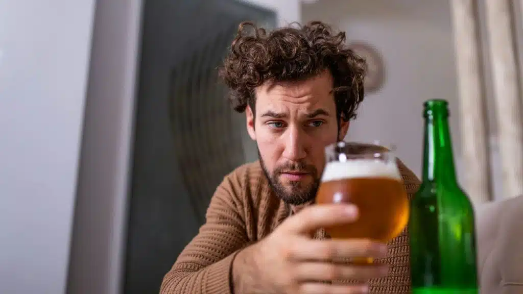 A man with curly hair looks at a glass of beer he is holding with a bottle next to it.