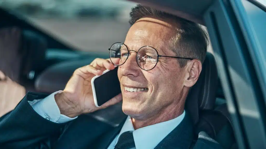A man in a suit and glasses is smiling while talking on a cell phone in his car.