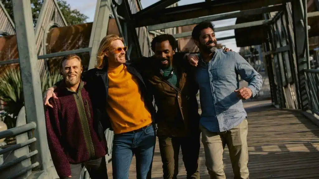 Four men walk across an industrial-style bridge with their arms around each other.