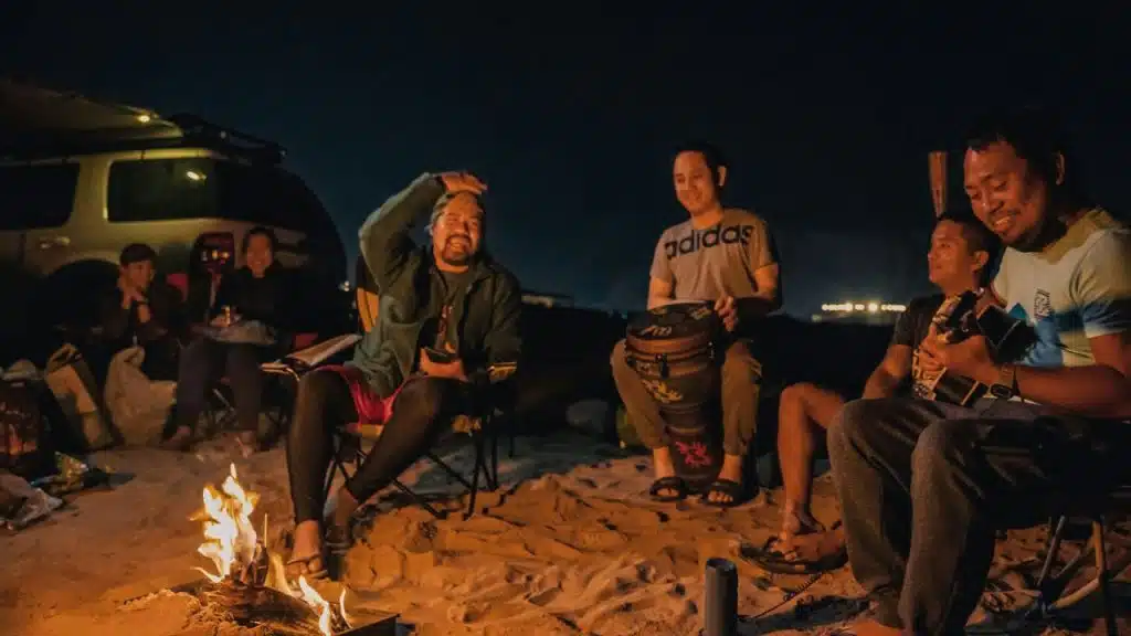 A group of men sit around a campfire on a beach at night, laughing and playing music.