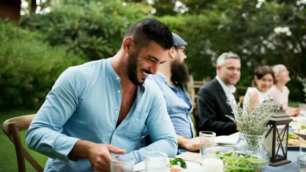 A bearded man in a blue shirt smiles and laughs with others at an outdoor table.