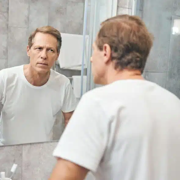 A man in a white t-shirt looks at his reflection in a bathroom mirror.