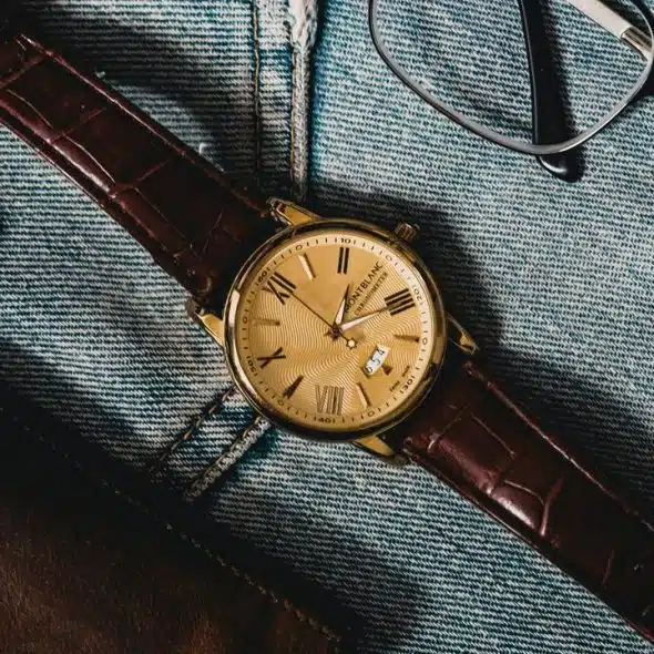 A gold wristwatch with a brown leather strap resting on denim next to eyeglasses.