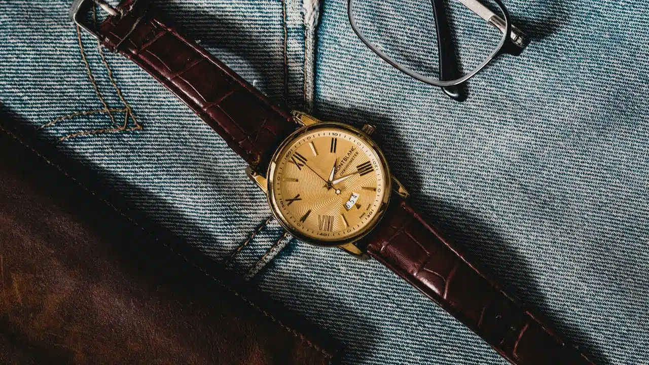 A gold wristwatch with a brown leather strap resting on denim next to eyeglasses.