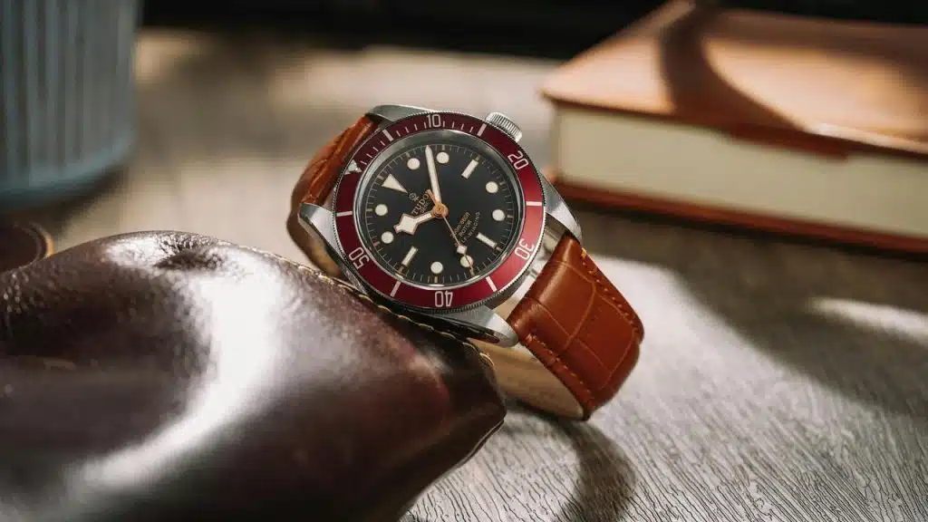 A Tudor dive watch with a red bezel and brown leather strap resting on a tabletop.