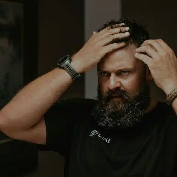 A worried man with a beard is looking in a mirror.