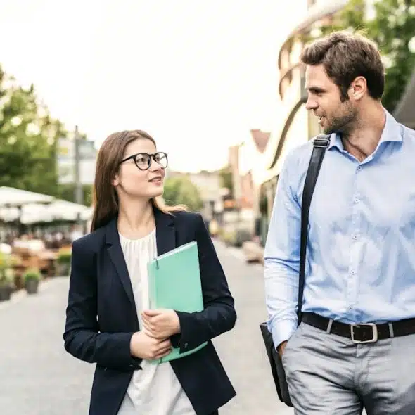 A woman wearing glasses and holding files in her hand walking down a street with a lightly bearded man who is carrying his bag on his shoulder and has his hands in his pockets.