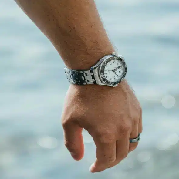 A silver dive watch with a white dial on a man's wrist by the water.