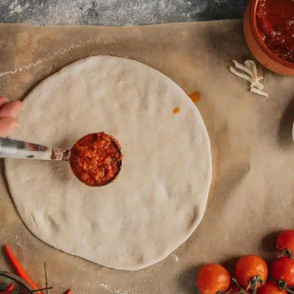 An overhead view of a person spreading tomato sauce on pizza dough.