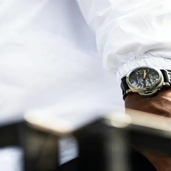 A close-up of a wrist wearing a Panerai watch with a black strap and a white jacket sleeve..