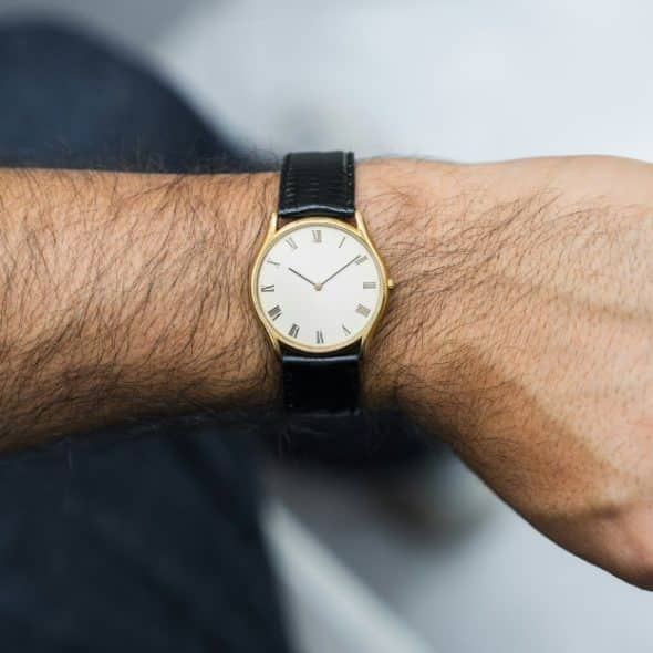 A man's forearm with a gold-tone watch with a white dial and black leather strap, positioned to show the watch clearly on the wrist.