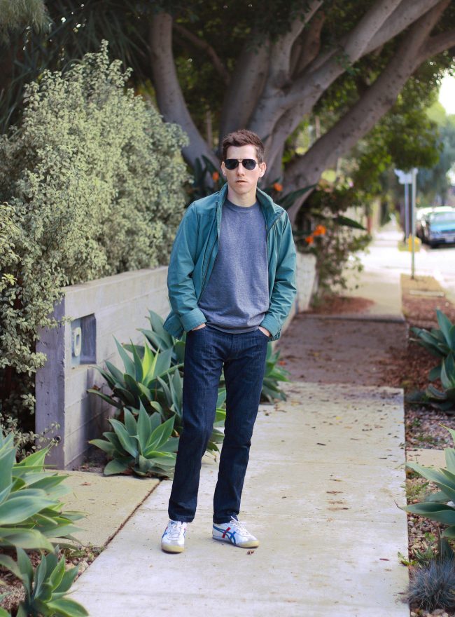 A man standing on a sidewalk in an outdoor setting, wearing a grey shirt, a green jacket, and dark wash jeans.