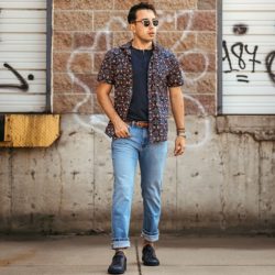 A man wearing light wash straight fit jeans with cuffs, a dark t-shirt, and a patterned short-sleeved shirt, standing in front of a brick wall.