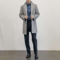 Man wearing a light gray overcoat, a dark blue dress shirt, black trousers, and black tassel loafers, standing against a white wall.