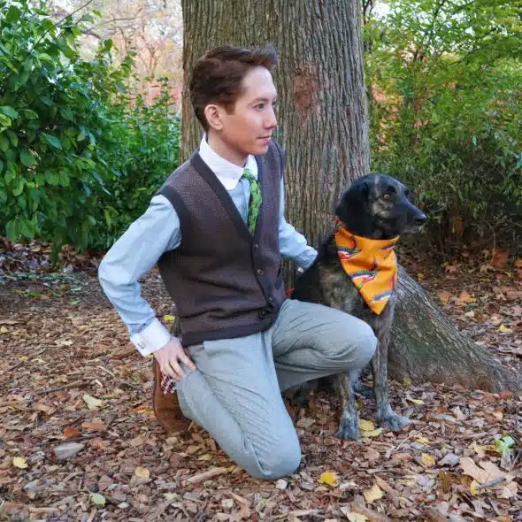A man kneeling next to a dog in a wooded area, wearing a vest, collared shirt, and tie.
