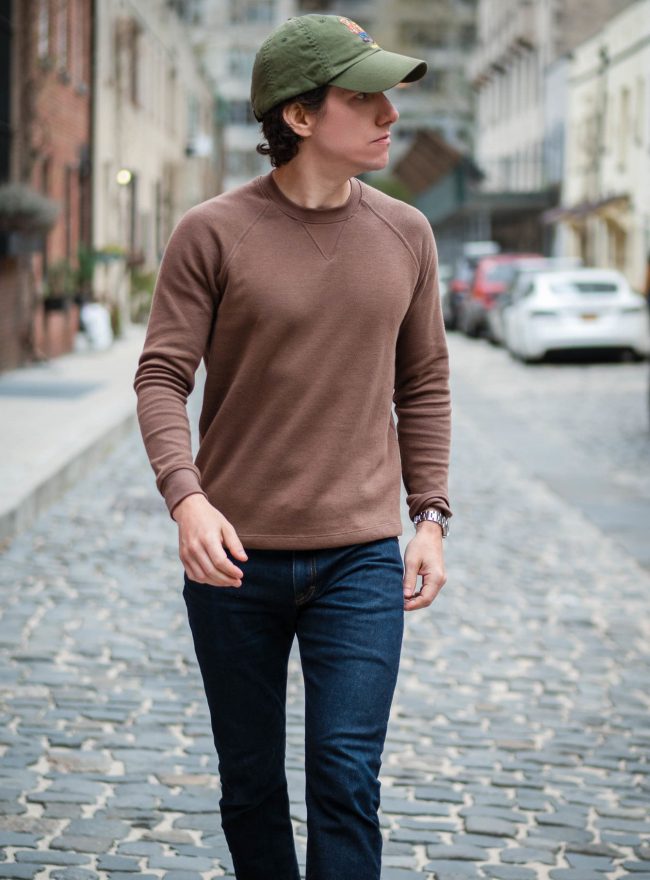 A man walking on a cobblestone street, wearing a brown sweater and dark wash jeans.