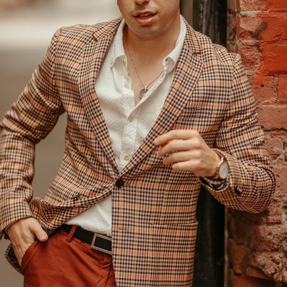 A close-up of a man wearing an orange plaid jacket over a white shirt and reddish-brown trousers.