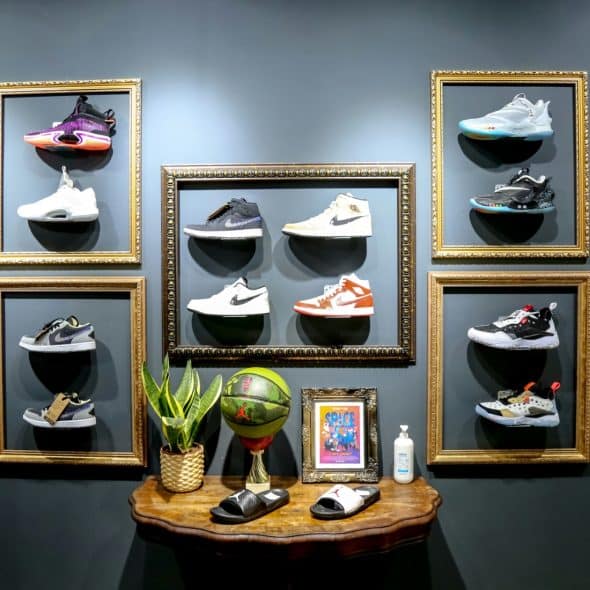 A collection of sneakers displayed in gold frames on a dark grey wall, with a small table below holding a basketball, plants, and other items.