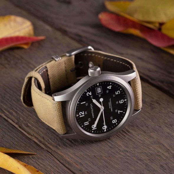 A Hamilton Khaki Mechanical watch with a black dial, Arabic numerals, and a beige canvas strap, lying on a wooden surface surrounded by yellow and red autumn leaves.