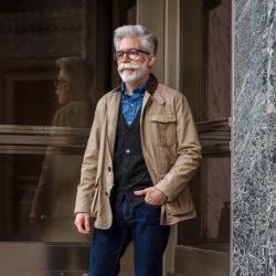 A man with a white beard and hair, wearing glasses, a tan utility jacket, a dark vest, a blue plaid shirt, and dark jeans, stands outdoors near a building entrance.