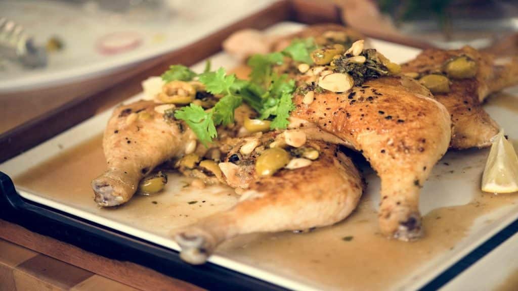 A plate of roasted chicken legs garnished with green olives, herbs, and nuts, served with a savory sauce and lemon wedges.