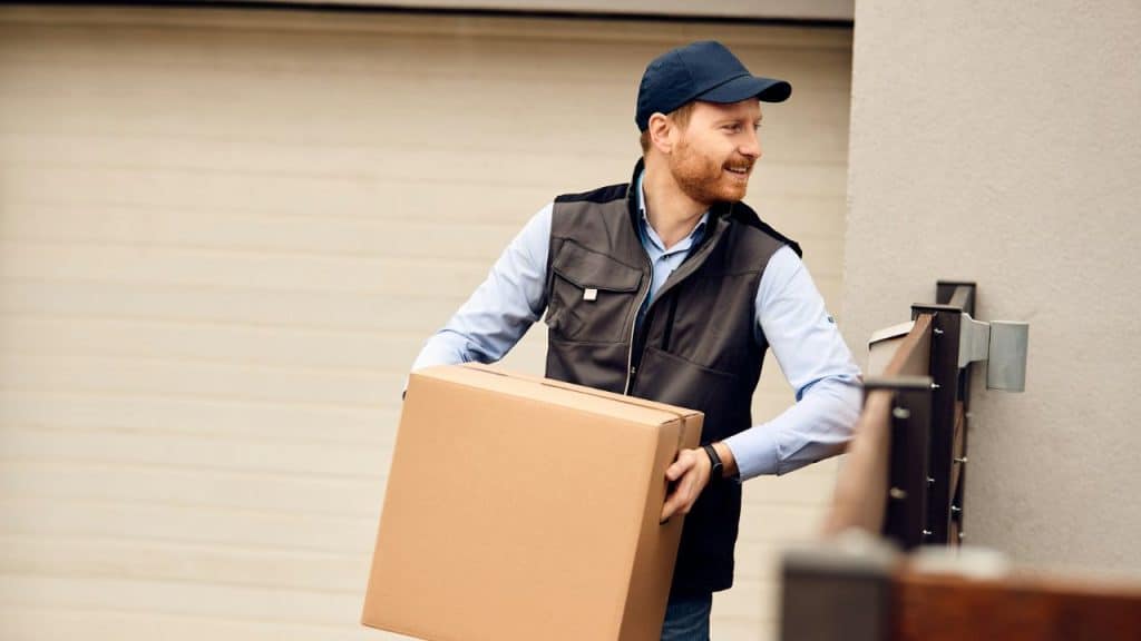 A smiling man in a vest and cap is carrying a large box toward a doorway.