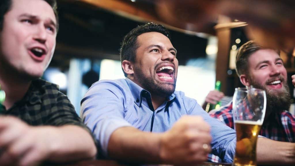 Three men at a bar are cheering with excited expressions and holding up their fists.