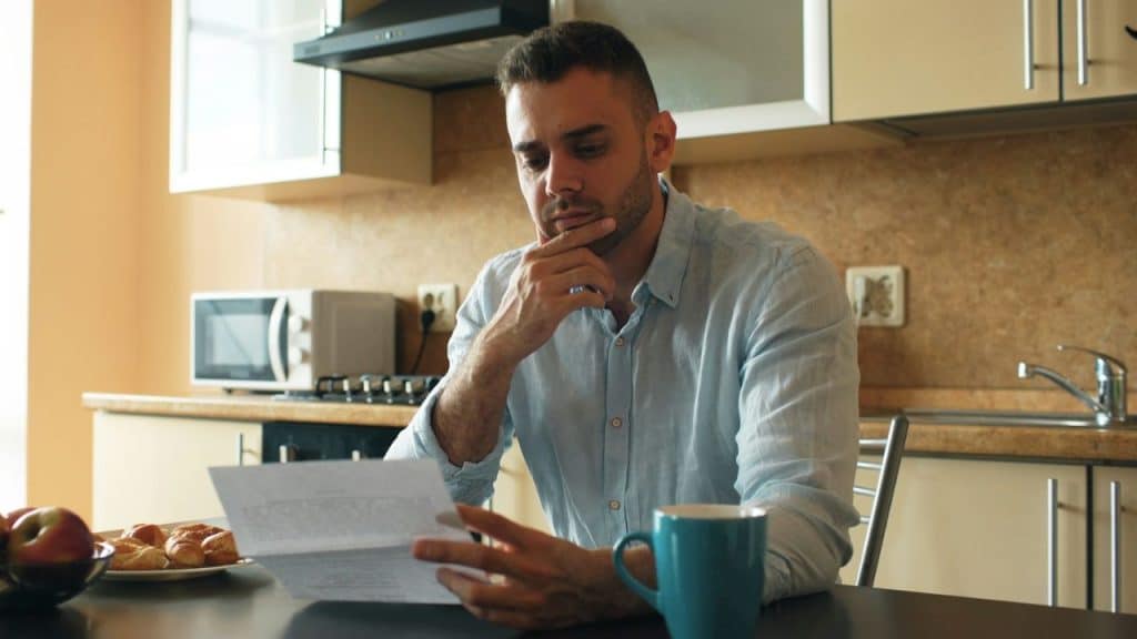 A man sits in a kitchen, holding a paper and resting his chin on his hand with a worried expression.