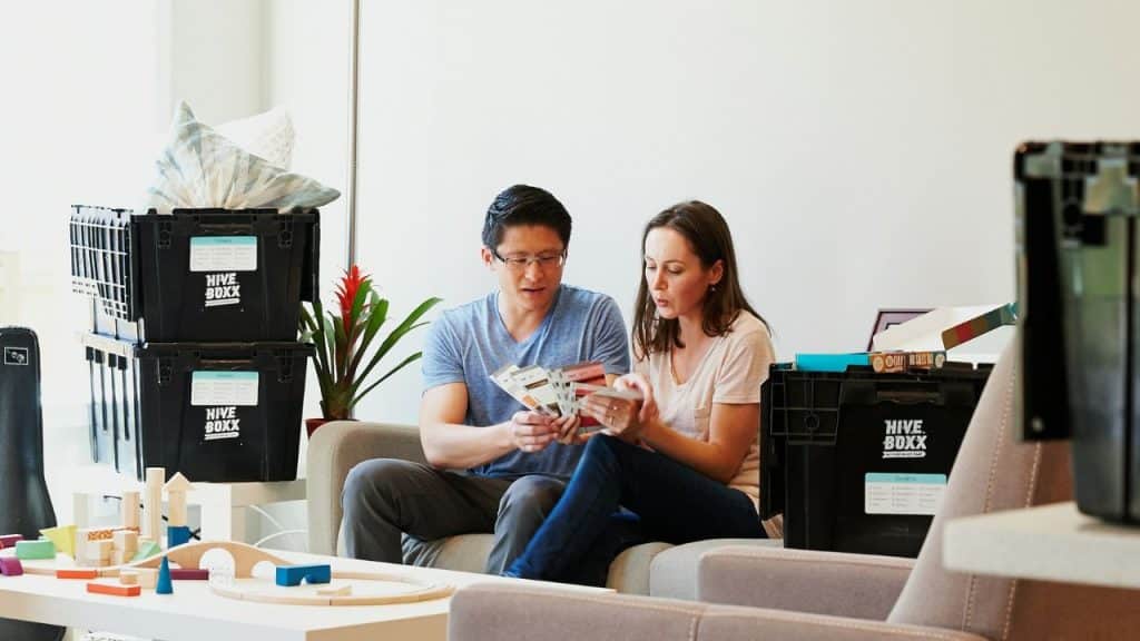 A man and a woman sit on a couch, looking at papers with moving boxes nearby.
