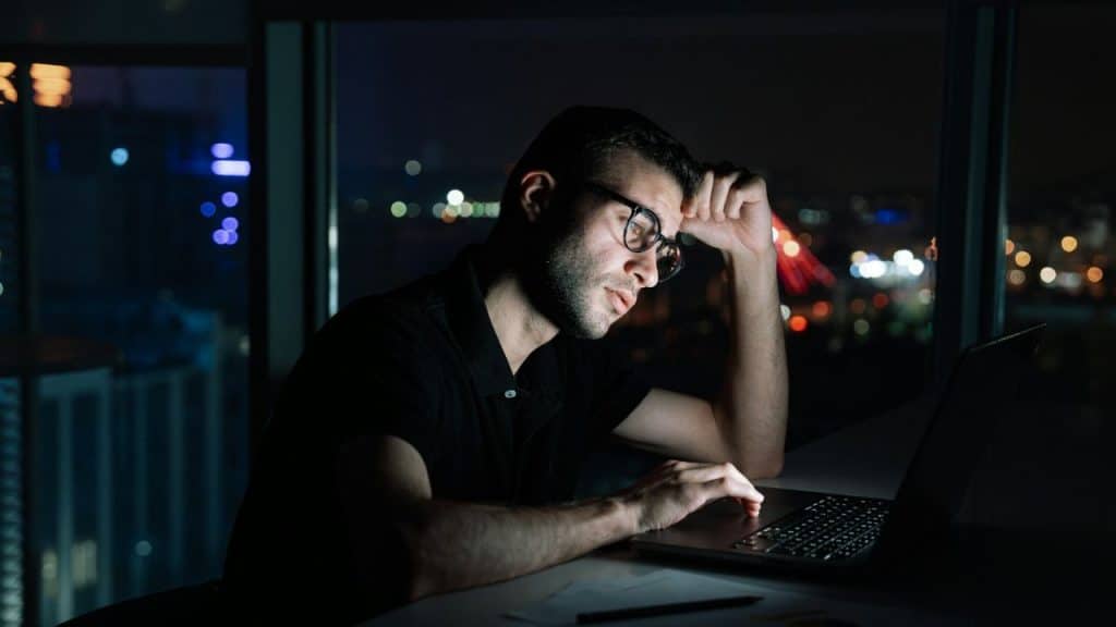 A man wearing glasses works on a laptop in a dark room with a city view at night.