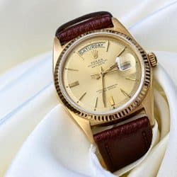 An elegant gold Rolex Day-Date watch with a champagne dial and brown leather strap, resting on white satin fabric.