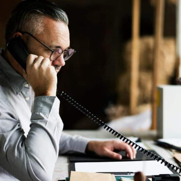 A man wearing glasses talks on a corded phone while working at a desk.
