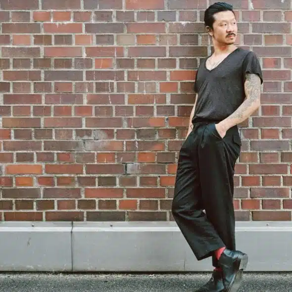 A man in a dark shirt and black pants leans against a brick wall.