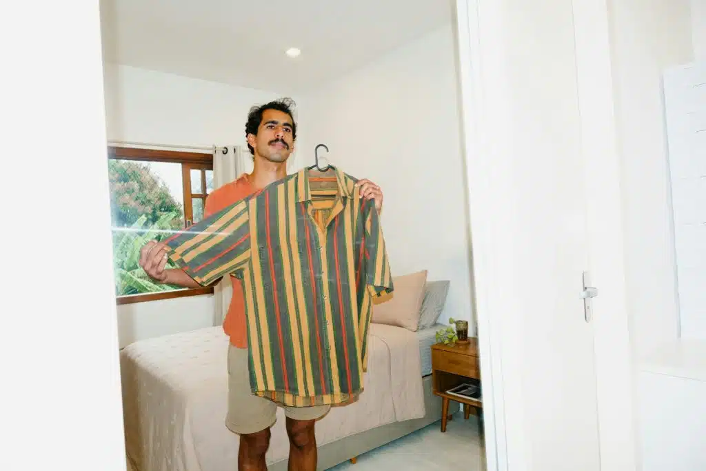 The man holds up the shirt while looking at the mirror.