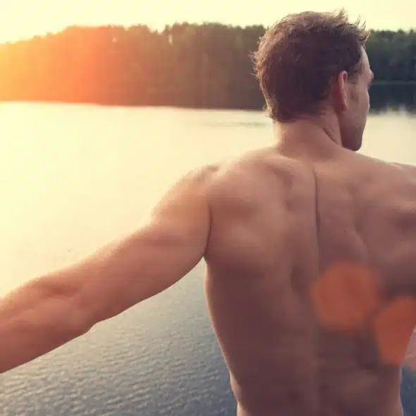 A muscular, shirtless man standing in front of a lake.
