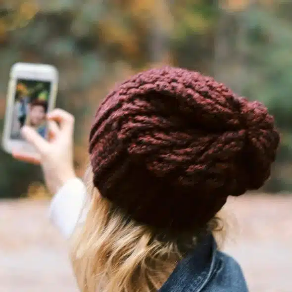 A person in a knit hat and denim jacket taking a selfie in an autumn forest.