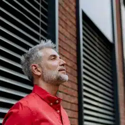 A gray-haired man in a red shirt looking upward beside a modern building.