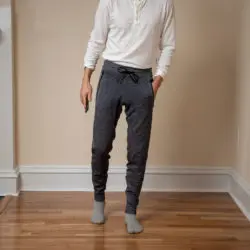 A person wearing a white long-sleeved shirt, gray joggers, and gray socks is standing on a wooden floor indoors.
