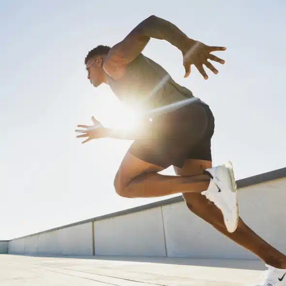 A man with dark skin and a muscular build is sprinting on a concrete surface under a bright, clear sky.