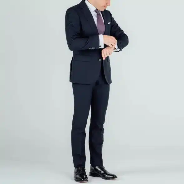 A man wearing a black suit with black oxford dress shoes looking at his wristwatch.