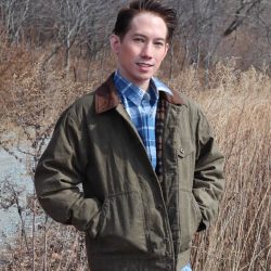 A man wearing a waxed jacket and plaid shirt standing in a field with tall grasses.