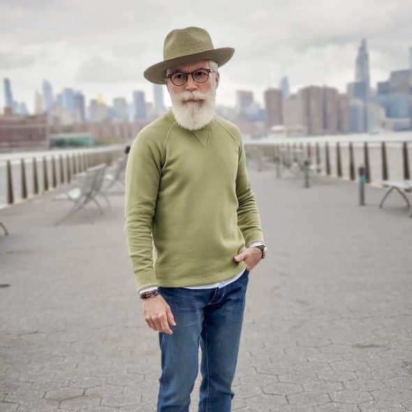 A man with a white beard wearing a hat, olive green sweatshirt, and relaxed fit jeans, standing outdoors with a city skyline in the background.