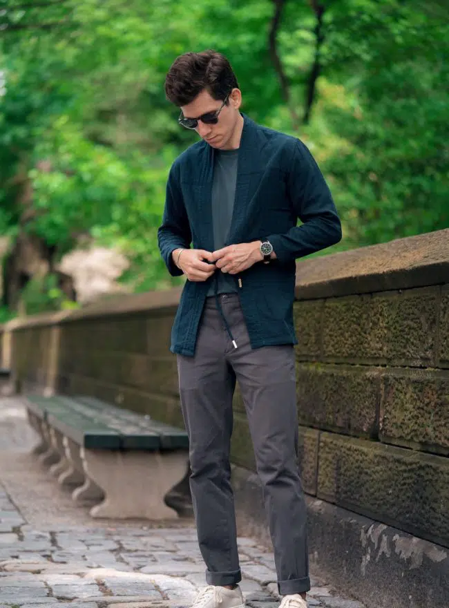 A man wearing sunglasses, a dark green Noragi jacket, a dark t-shirt, and gray pants with white sneakers, standing on a stone pathway outdoors