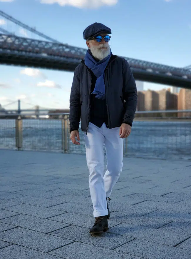 A man walking on a paved area, wearing a dark jacket, a blue scarf, a blue cap, white pants, and dark boots, with a bridge in the background.
