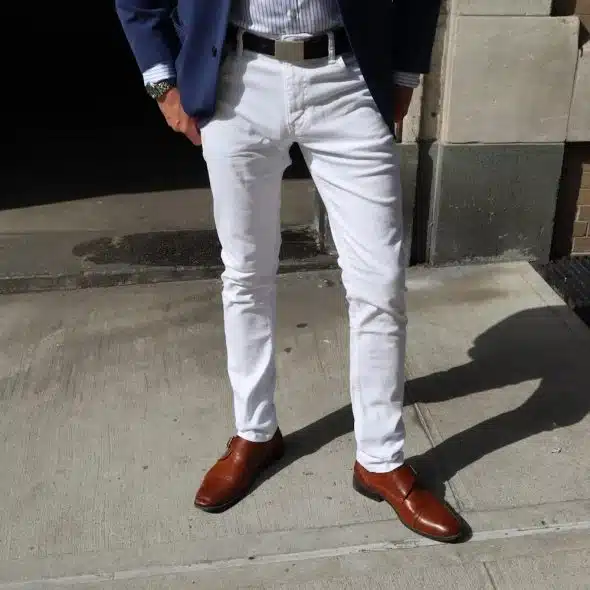 A man's lower body wearing white jeans and brown monk strap shoes, with a striped shirt and dark jacket visible.