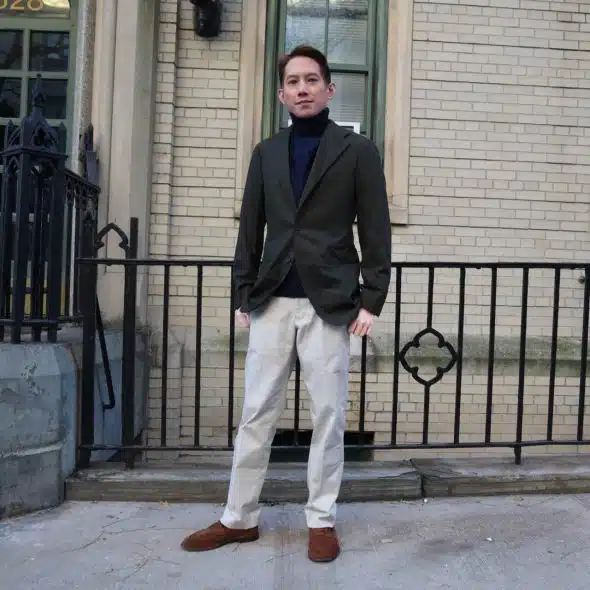 A man wearing a dark blazer, turtleneck, and light-colored pants standing in front of a brick building.