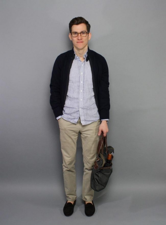 Man in a striped casual button-up shirt, dark cardigan, and chinos holding a bag against a grey background.