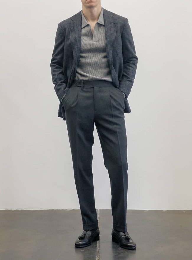 A man wearing a gray plaid suit jacket over a light gray knitted polo shirt, with matching gray trousers and black dress shoes, standing against a white wall.