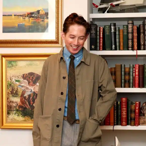 A man smiling and wearing a light brown jacket, blue shirt, and tie, standing in front of bookshelves.