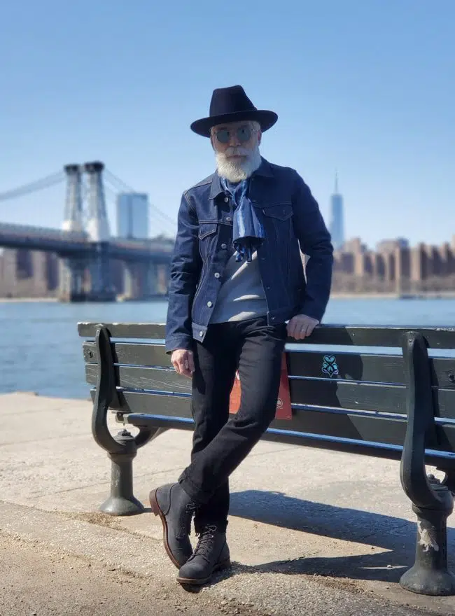 An older man with a white beard wearing a dark hat, denim jacket, grey shirt, black jeans, and dark boots, leaning on a park bench by a body of water with a bridge in the background.